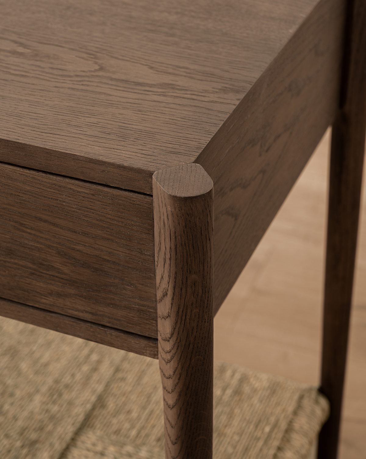 Close-up of the Dana 2-Drawer Console’s oak corner with rounded legs and visible wood grain, set above a woven mat on a light wood floor—ideal for adding style to any entryway, by McGee & Co.