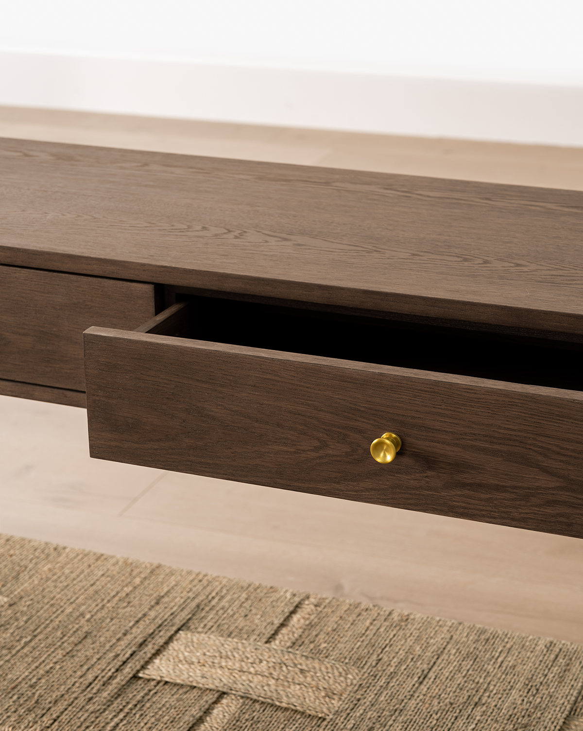 The Dana 2-Drawer Console in dark oak features a partially open drawer with a round gold knob, set on a light wood floor and woven beige rug—an elegant addition to entryway decor, by McGee & Co.