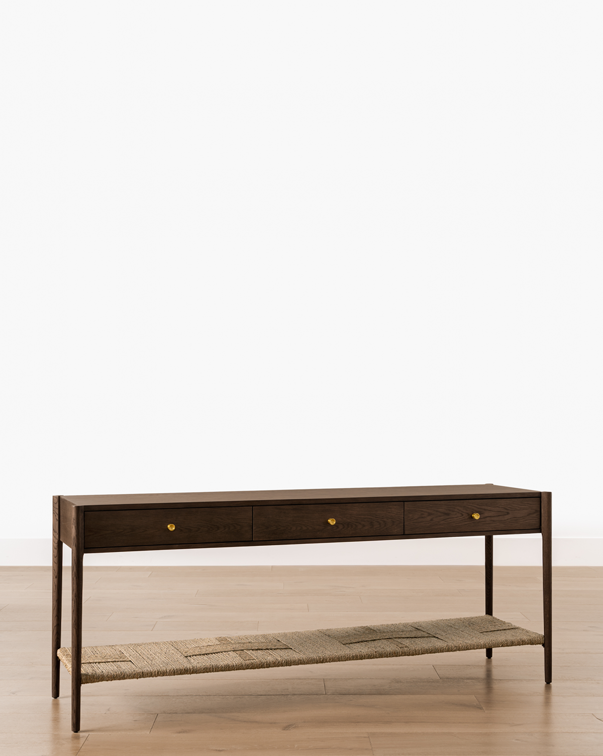 The Dana Walnut Oak Console features three small drawers with brass hardware and an open lower woven shelf, shown on a light wood floor against a plain white background, by McGee & Co.