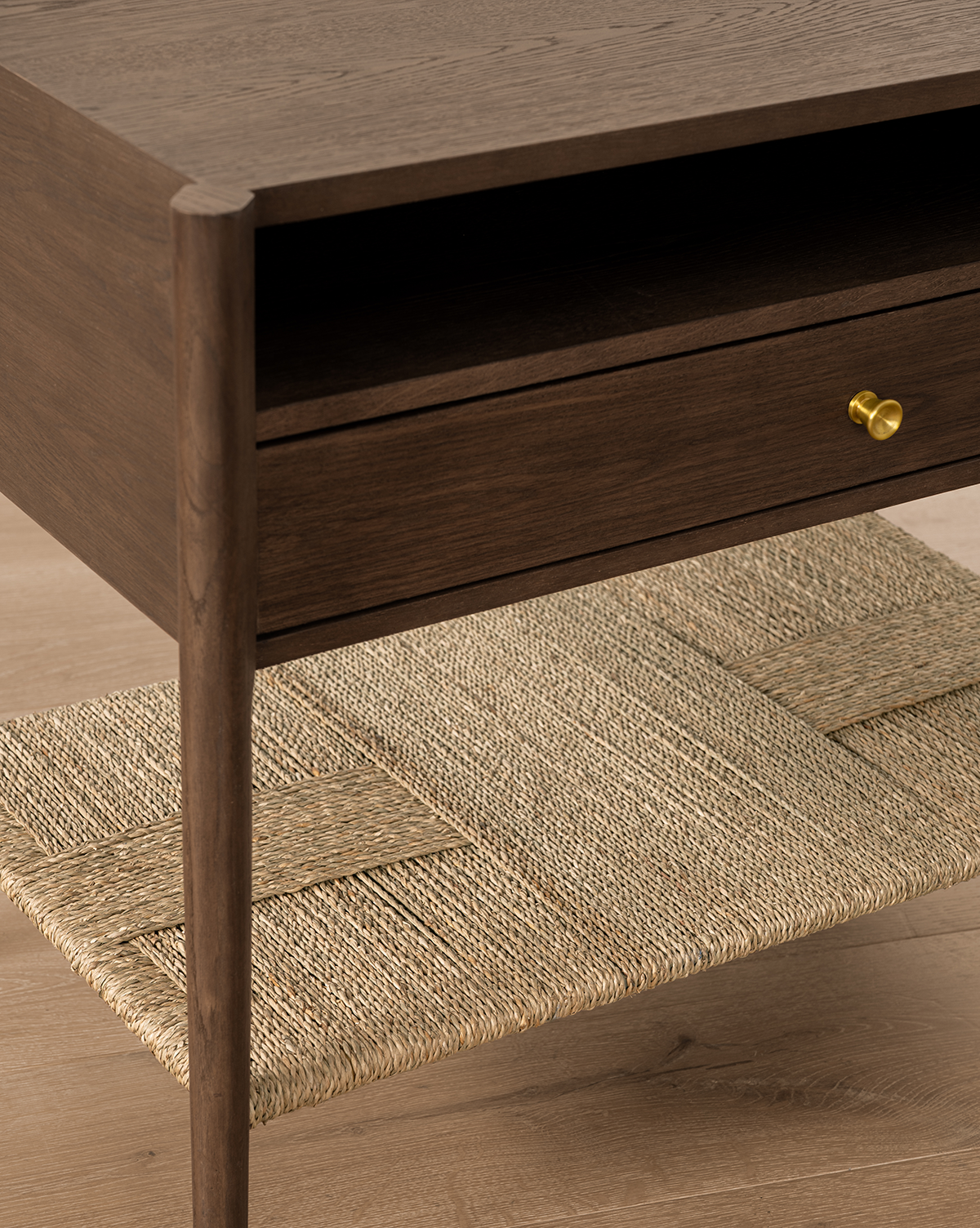 A close-up of the Dana Walnut Oak Nightstand featuring a drawer with brass hardware and an open rattan shelf, set on a light wood floor, by McGee & Co.