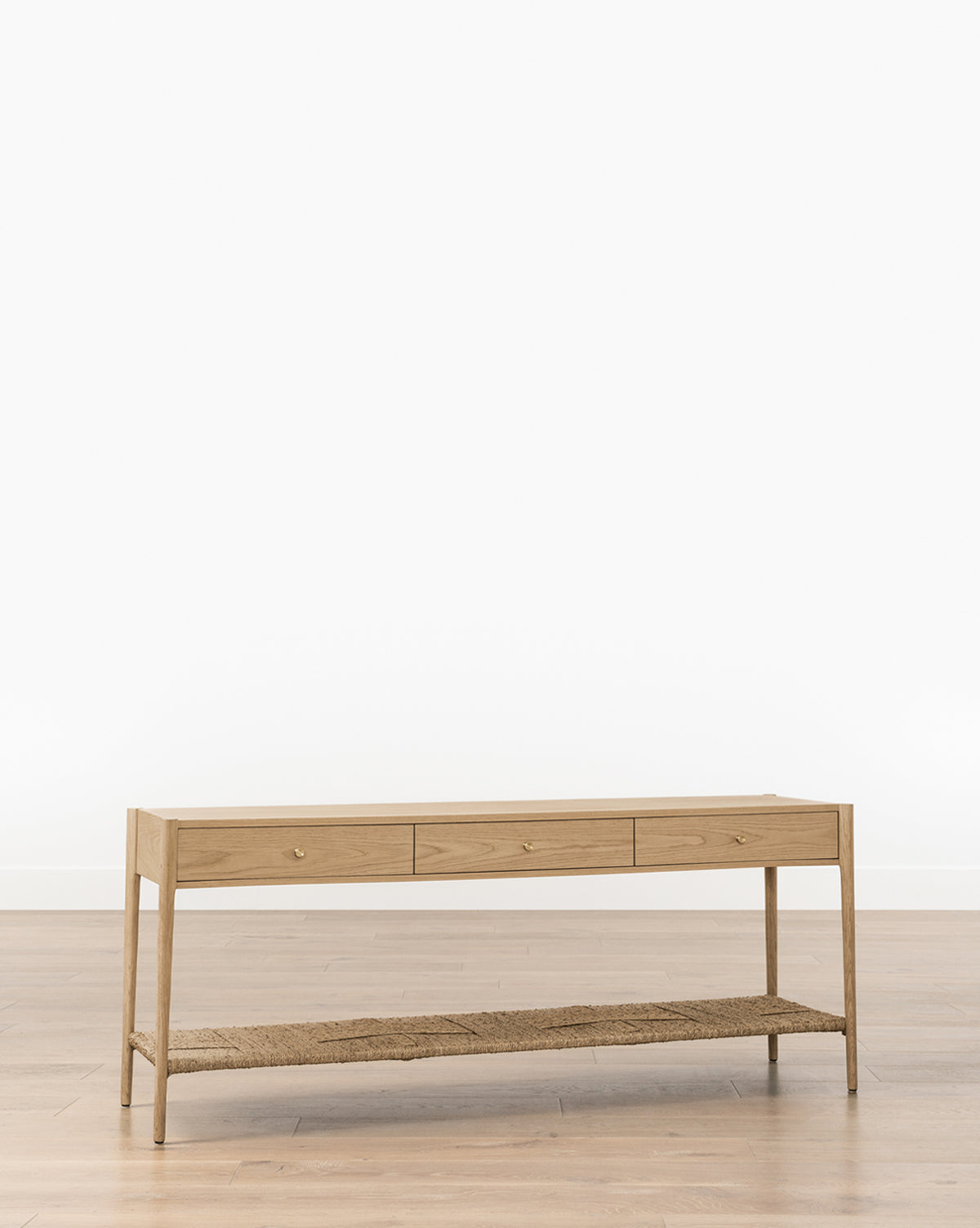 The Dana White Oak Console features a minimalist design with soft-close drawers and a woven seagrass shelf, showcased against a plain white wall on a light wood floor, by McGee & Co.