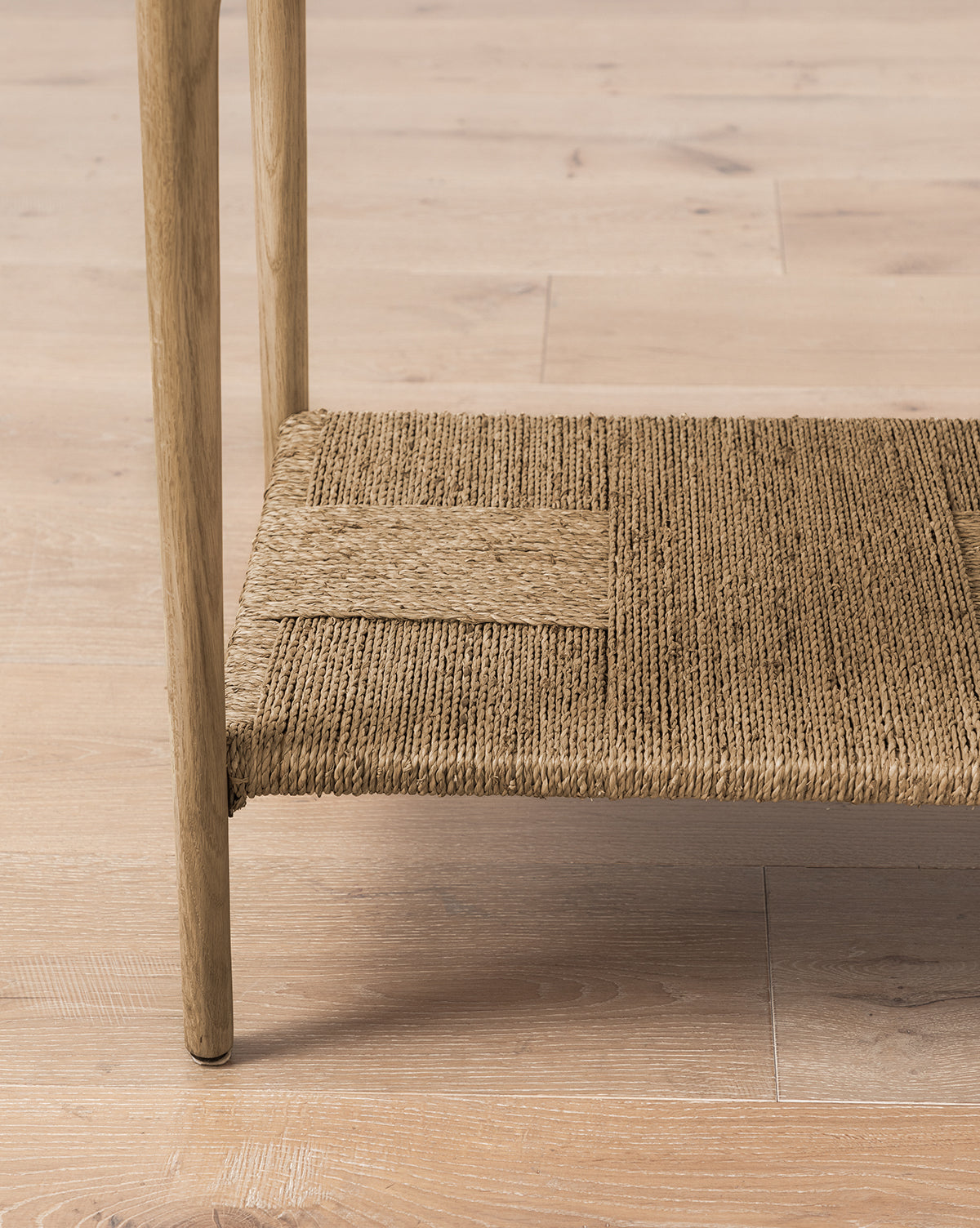 A close-up of the Dana White Oak Console’s leg and its woven seagrass shelf, featuring a natural textured weave, placed on light wood flooring by McGee & Co.