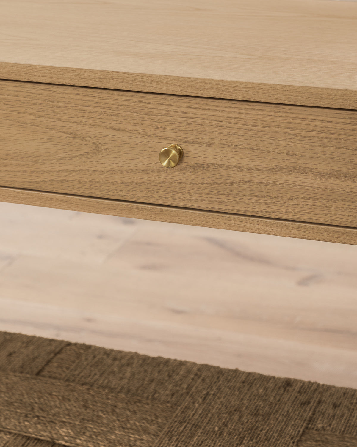 Close-up of the Dana White Oak Console’s wooden drawer with a round brass knob, shown above a textured woven seagrass shelf and matching wooden surface by McGee & Co.