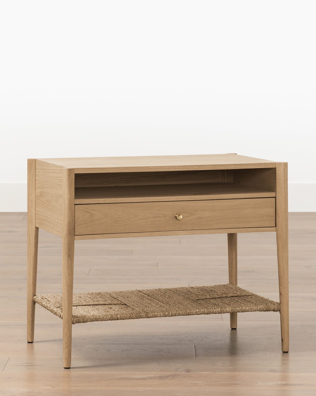 The Dana White Oak Nightstand features tapered legs, an open shelf, a single drawer with brass hardware, and a woven seagrass shelf, set on a wooden floor against a plain white background, by McGee & Co.