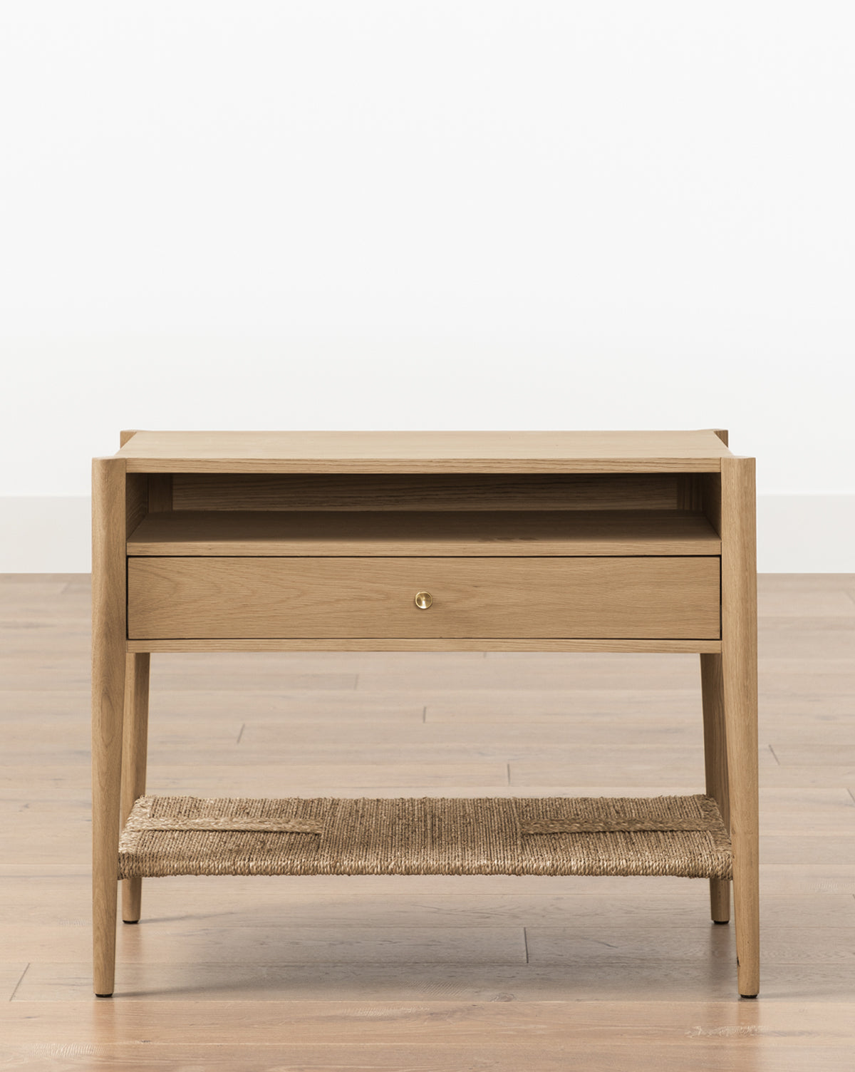 The Dana White Oak Nightstand features a single drawer with brass hardware, an open shelf, and a woven jute lower shelf, by McGee & Co. Its clean, modern lines and four tapered legs stand out against a plain white background.
