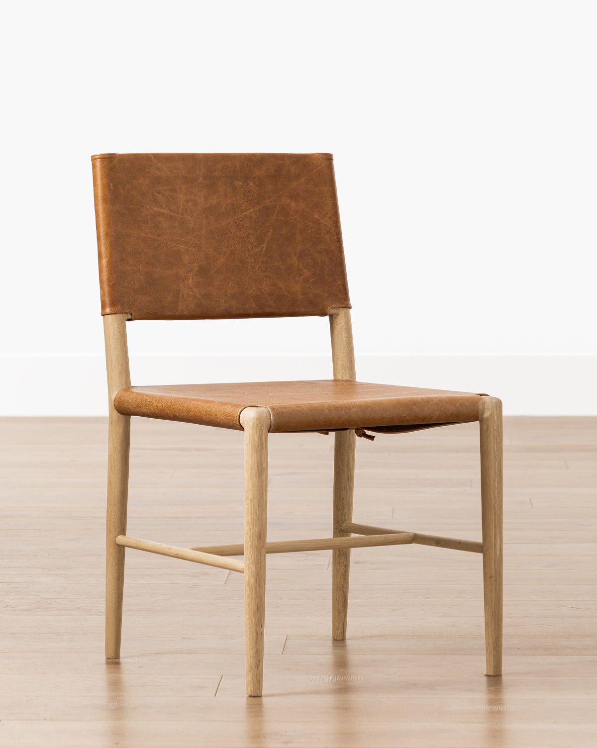 The Danson Dining Chair features a light wooden frame with brown leather seat and backrest, showcasing a minimalist design. It is shown on a wooden floor against a plain white wall by McGee & Co.