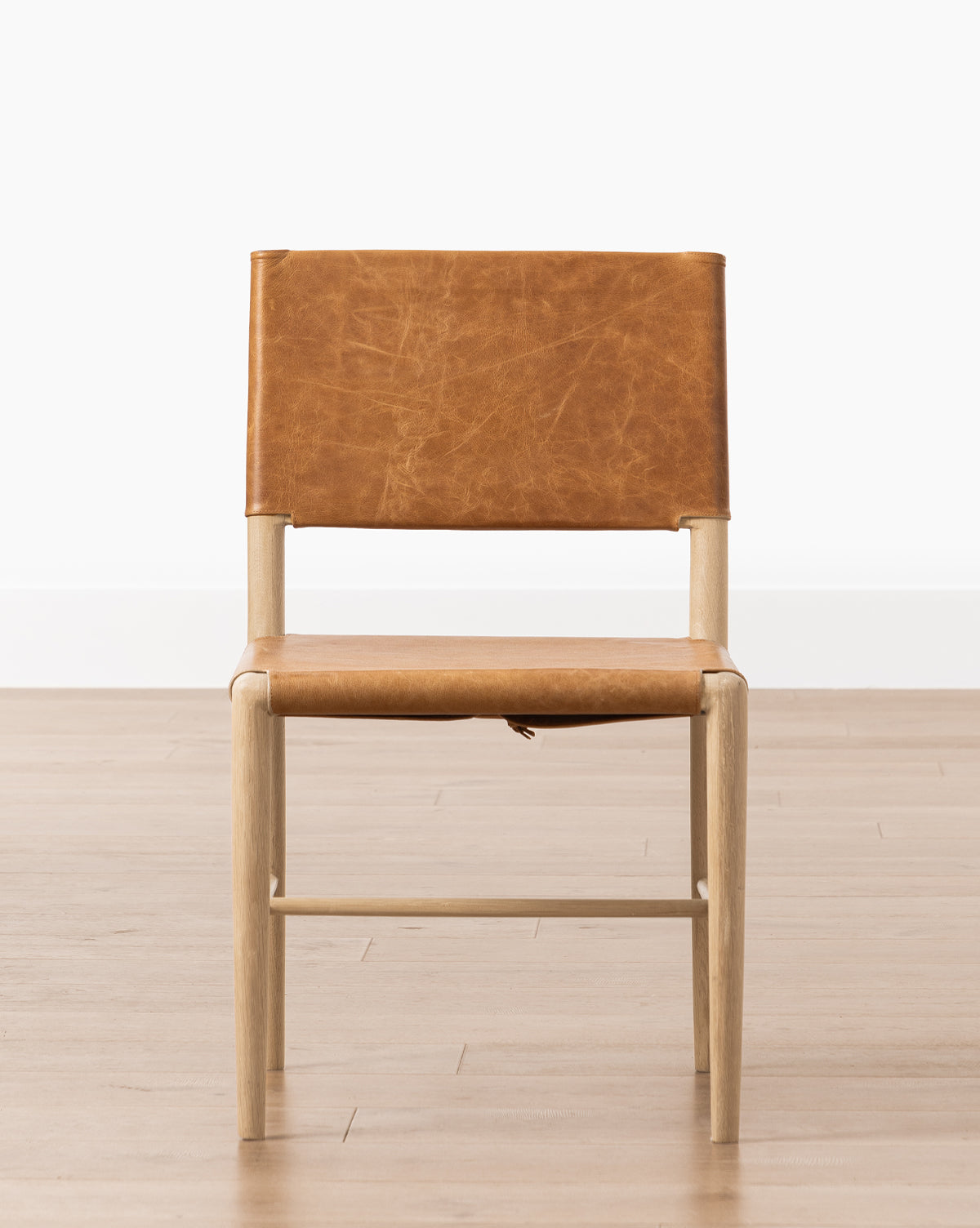 The Danson Dining Chair has a minimalist design with a light wood frame and tan leather seat and backrest, placed on a light wood floor against a plain white wall by McGee & Co.