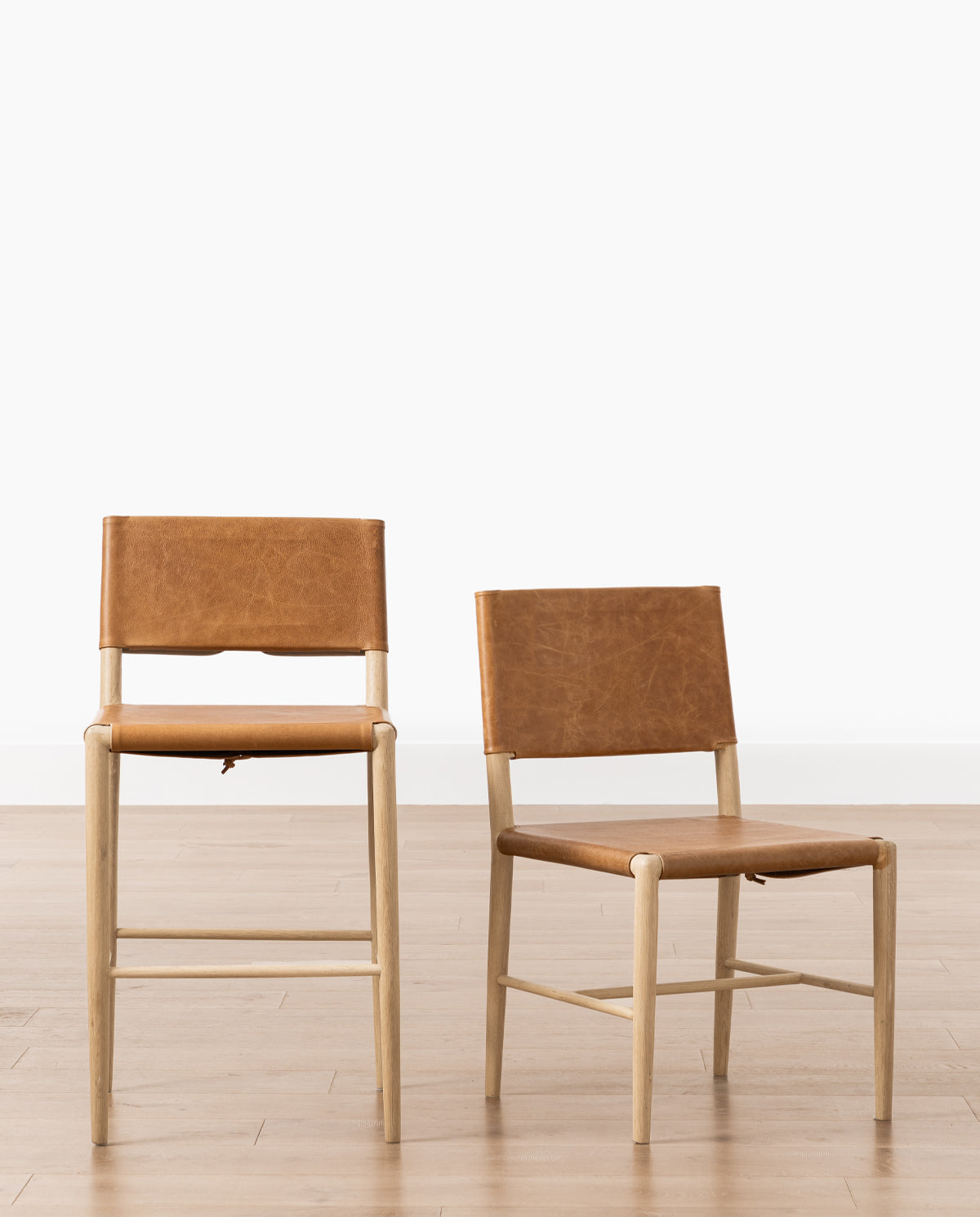 A Danson Dining Chair and a matching bar stool, both with minimalist wooden frames, light tan legs, and brown leather seat and back, are set on a wooden floor against a white background by McGee & Co.
