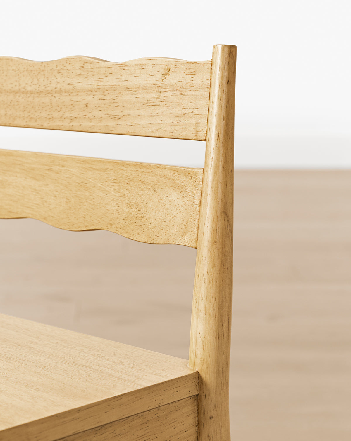 Close-up of the backrest and seat of a Darcy Kids Chair from the Darcy Kids Chairs (Set of 2), featuring a smooth, natural finish and a wavy top slat. The background shows a plain, light-colored floor and wall, by McGee & Co.