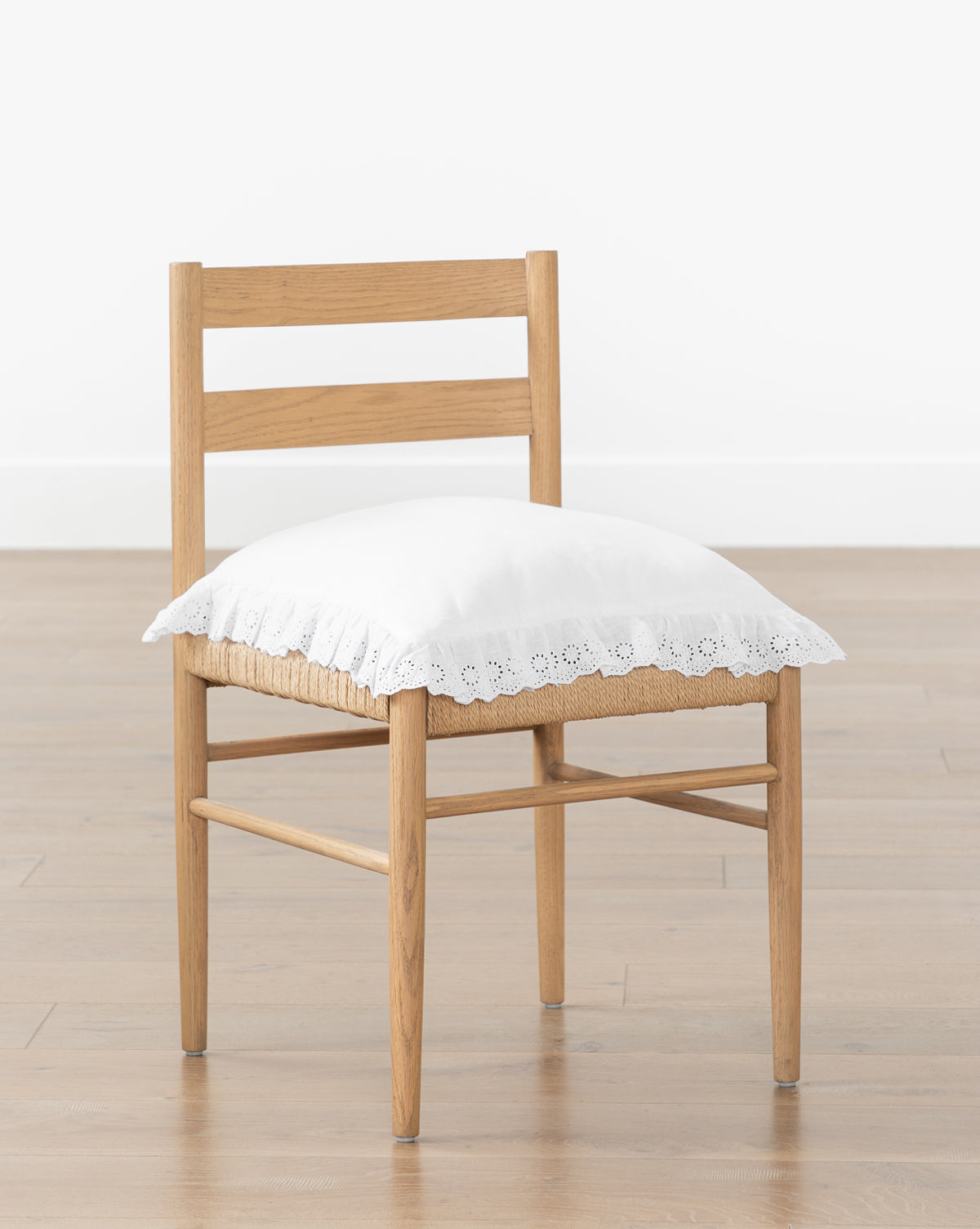 A wooden chair with a ladder back sits on a light wood floor against a white wall, topped with the McGee & Co. x Loeffler Randall Delilah Broderie Trim Seat Cushion from McGee & Co., blending timeless design and contemporary style from McGee & Co.