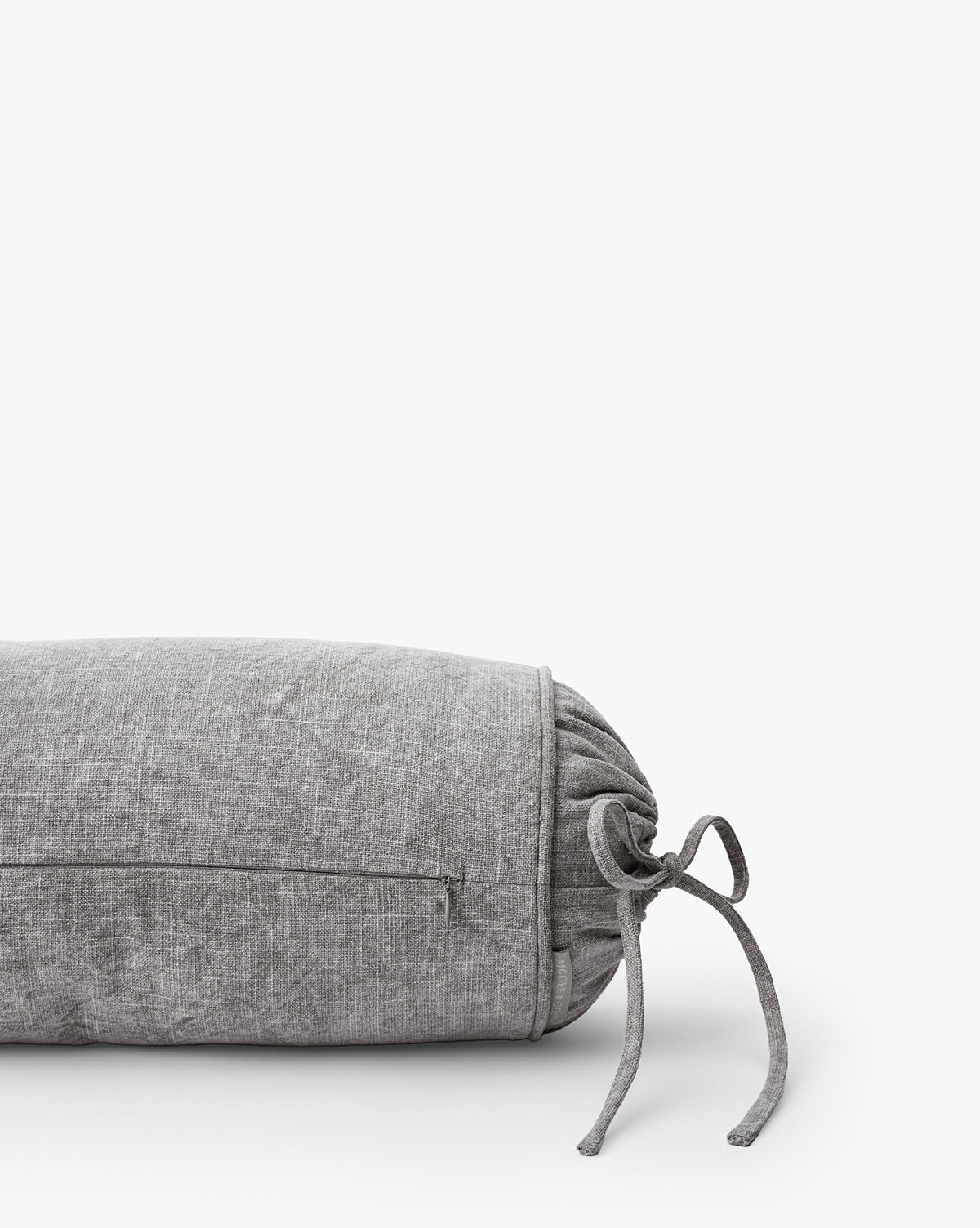 The Delphine Bolster Pillow is a vintage-inspired gray stonewashed cotton pillow with a fabric tie and side zipper pocket, shown against a plain white background.
