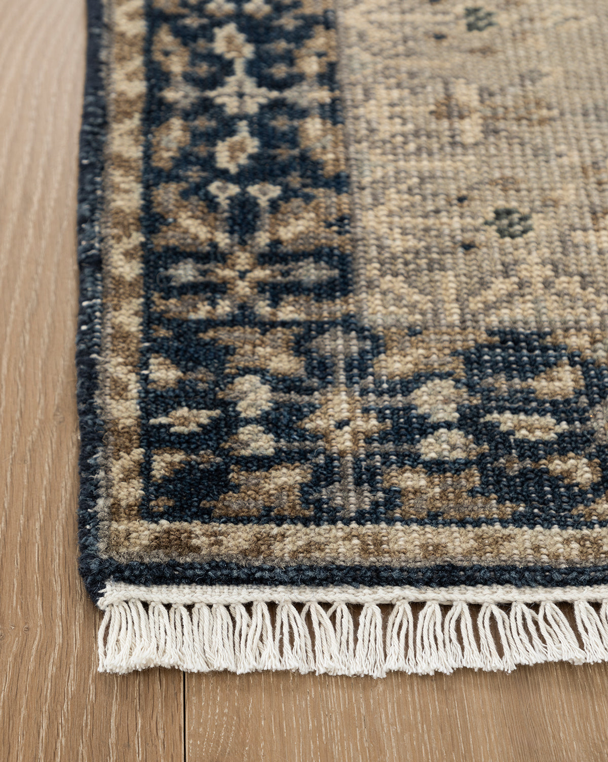 A close-up of the Deveraux Hand-Knotted Wool Rug corner on a wooden floor, showcasing beige, navy, and brown hues with fringe along the edge, by McGee & Co.