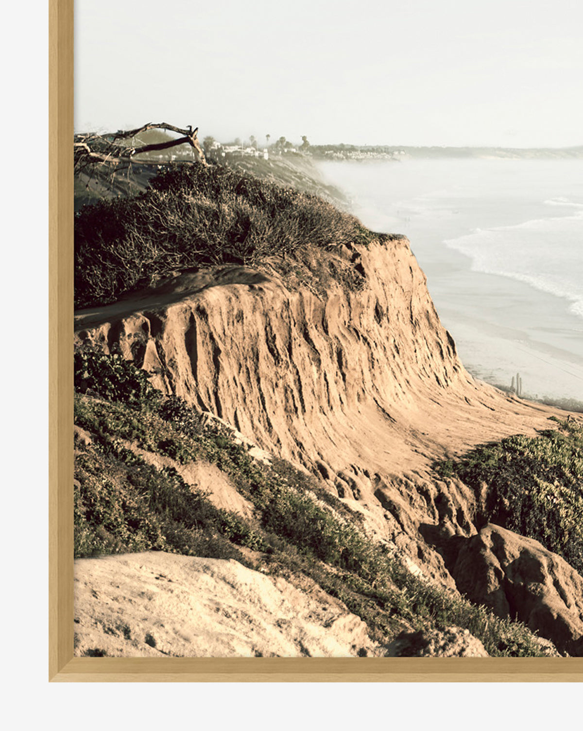 Distant Coastline: A giclee wall art print showcasing rugged sandy cliffs with green shrubs above a misty ocean beach, elegantly framed in light wood.