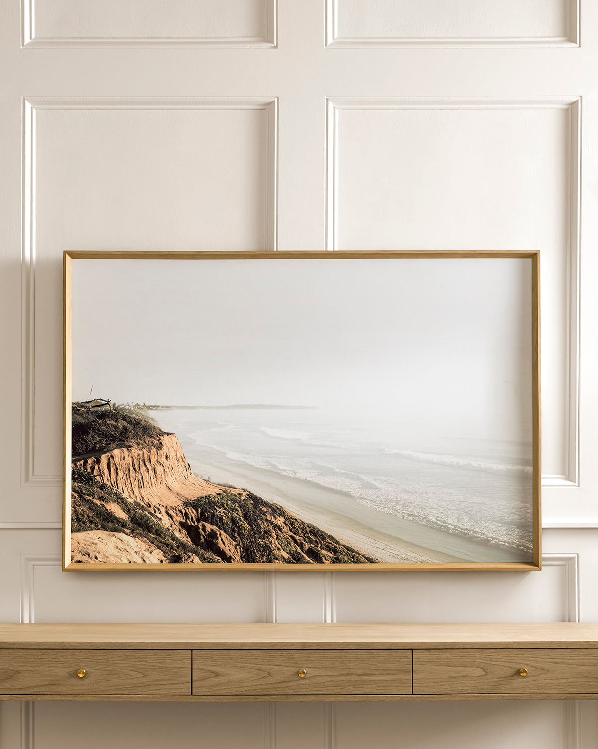 A framed Distant Coastline giclee art print of a misty shoreline hangs on a white paneled wall above a wooden shelf with drawers.