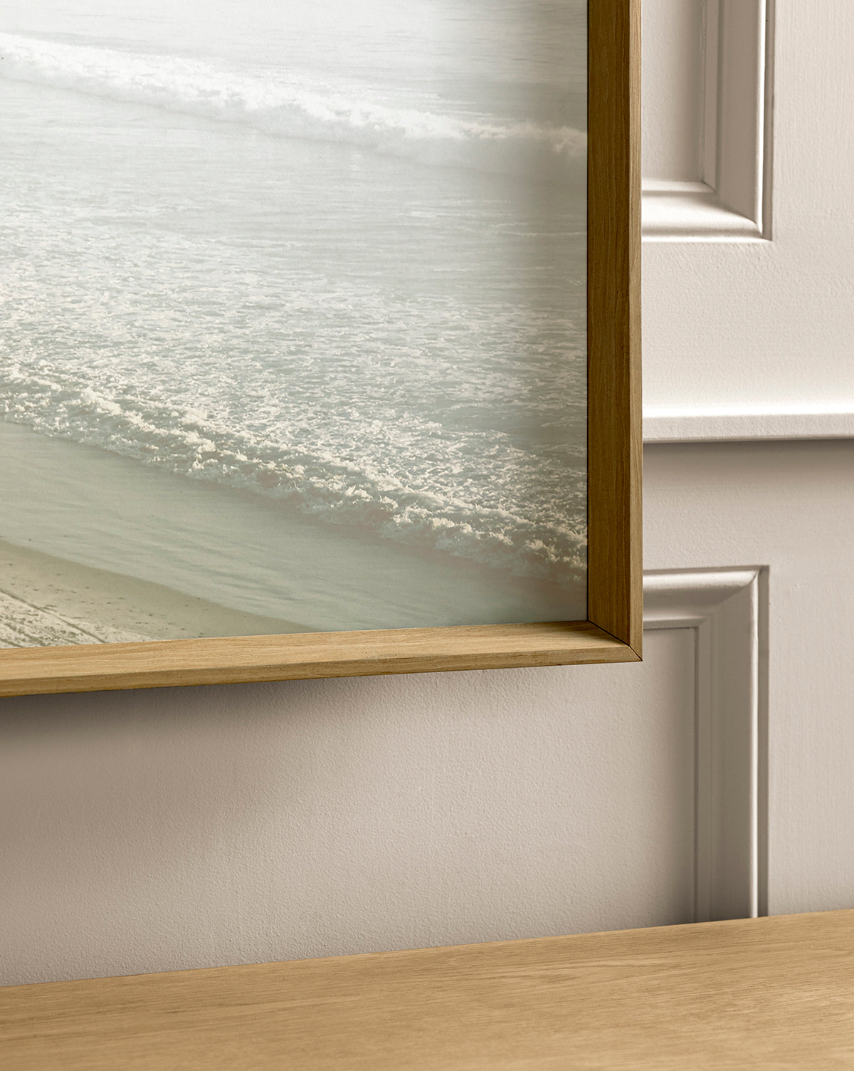 A close-up of the Distant Coastline coastal wall art: a framed giclee print showcasing ocean waves by the sandy shore, displayed on a light-paneled wall above a wooden surface.