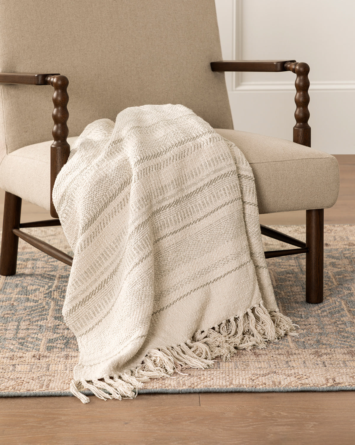 The Donahue Striped Throw, a cream-colored, hand-woven cotton blend with fringe, is draped over the arm of a beige upholstered chair with wooden arms, resting on a patterned rug in a cozy, well-lit room, by McGee & Co.