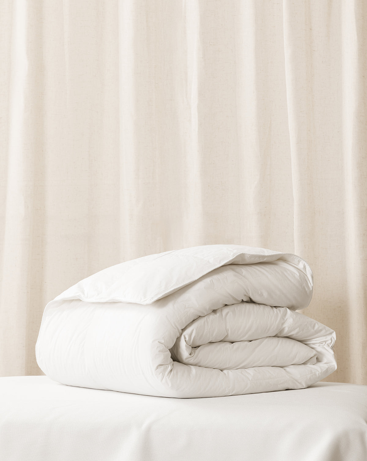 A folded white down duvet insert on a bed with a beige curtain in the background from McGee & Co.