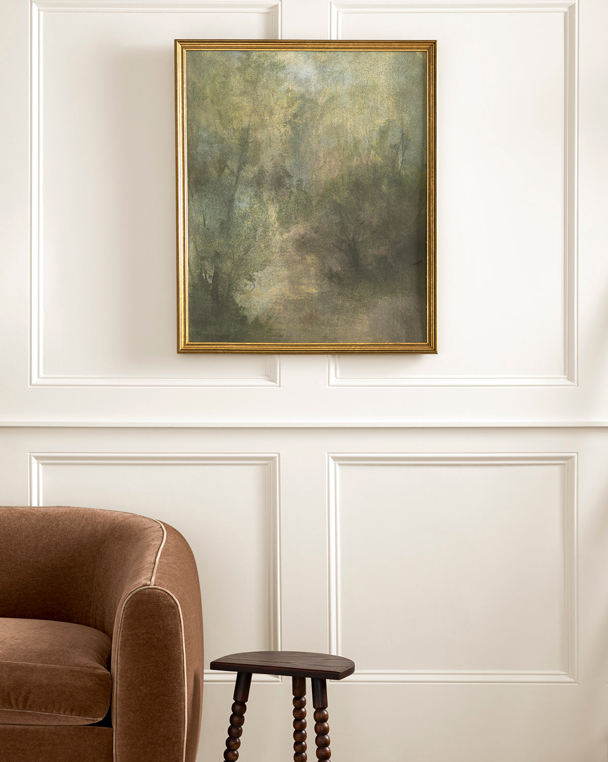 The framed Dream State landscape painting with soft green and brown tones hangs on a white paneled wall above a brown upholstered chair and a small dark wooden side table, by McGee & Co.