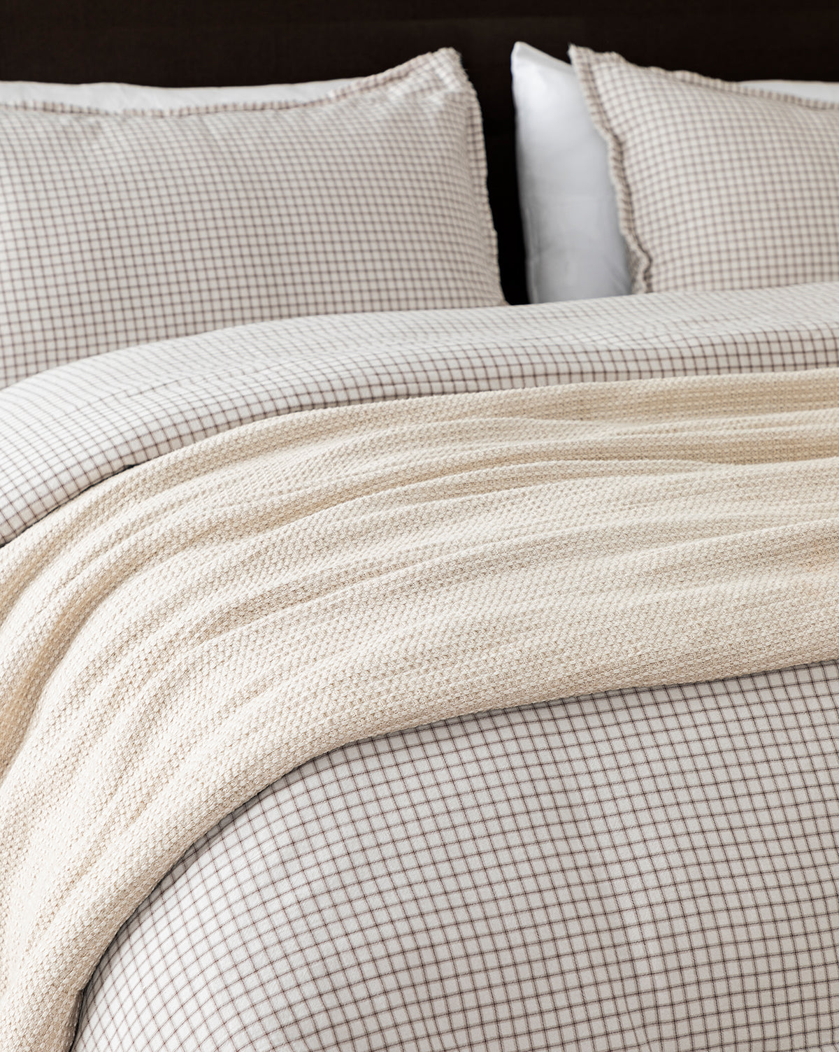 A neatly made bed with a beige and white small checkered duvet and matching pillows is topped with the Penedo Duffy Oversized Throw, a cream-colored knit blanket in neutral tones draped across the foot of the bed, McGee & Co.