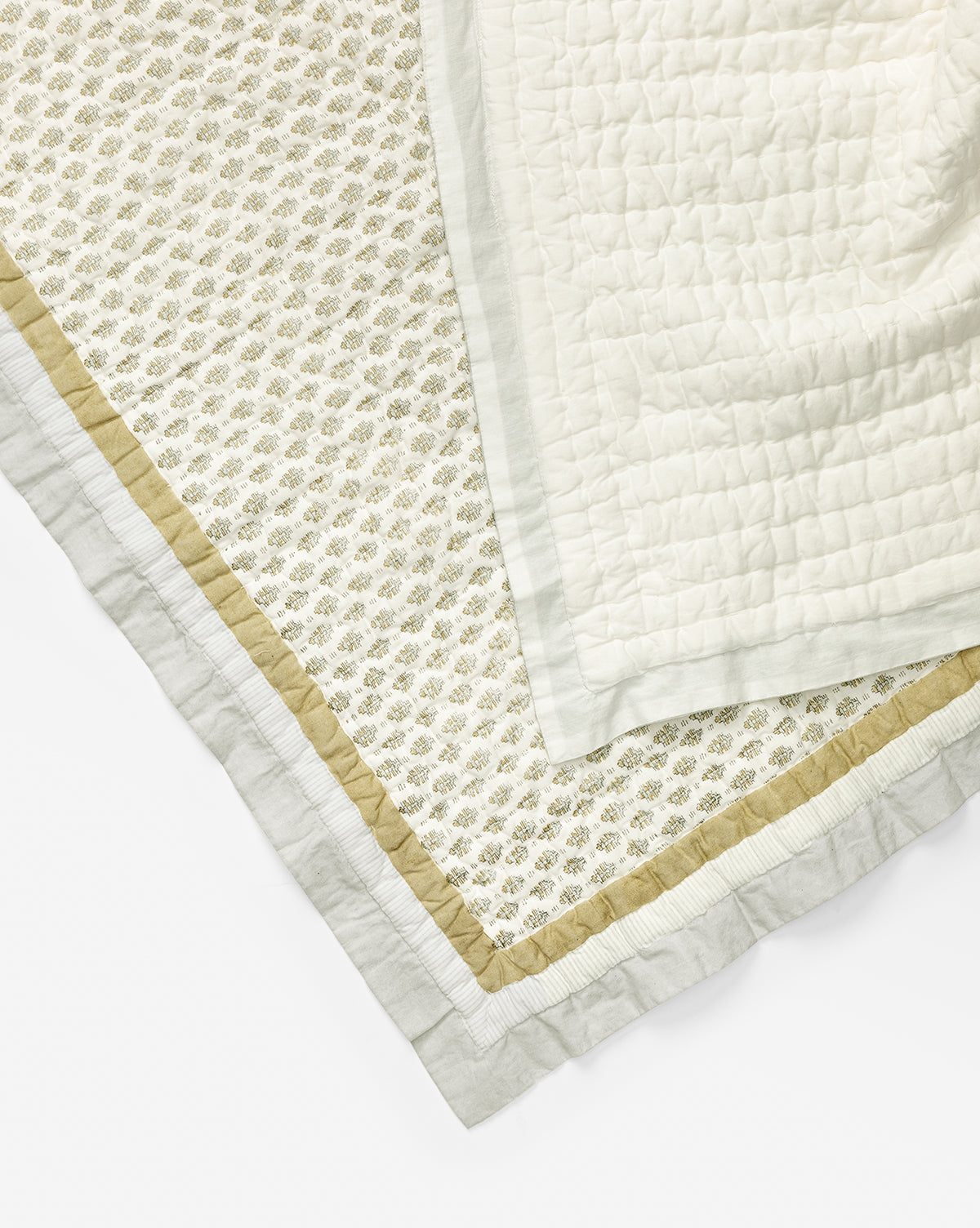 A close-up of two overlapping blankets: a cotton quilt with a quilted texture, and the Renuka Inc. Dulcie Block Print Kids Quilt with a small floral pattern and green and gold-toned borders, both in light, neutral colors—McGee & Co.
