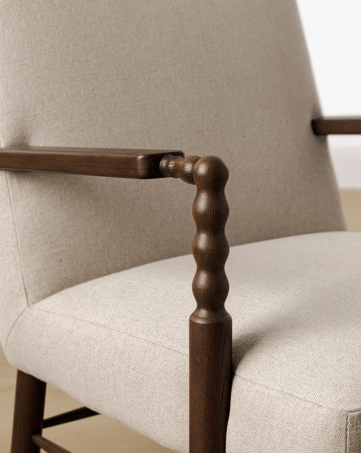 Close-up of the Earnshaw Lounge Chair showcasing neutral-toned, textured upholstery with dark wood armrests and legs in an English oak finish, by McGee & Co. Foreground displays a rounded spindle armrest design set against light-colored flooring and walls.