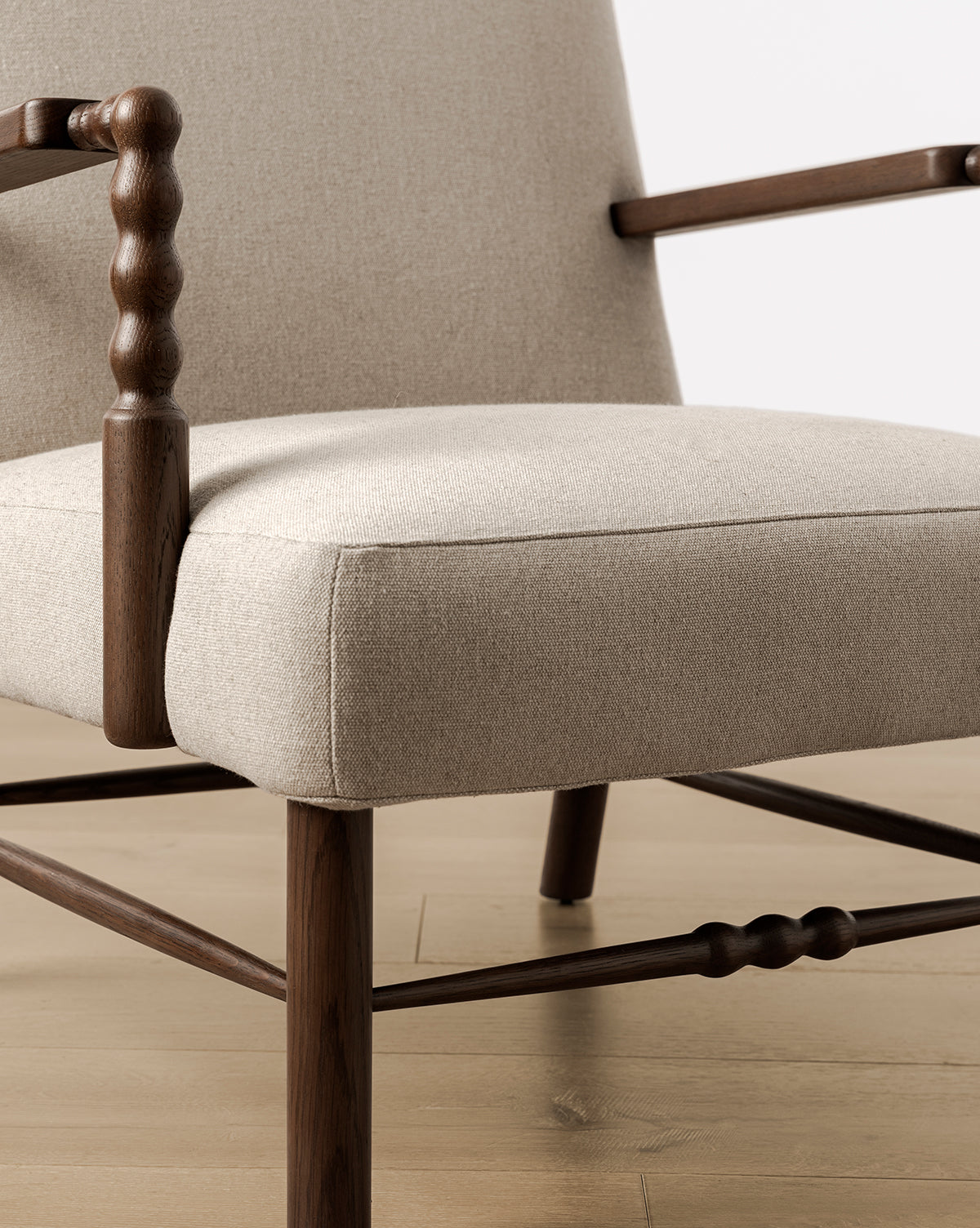 Close-up of the Earnshaw Lounge Chair, showcasing neutral upholstery and wooden armrests and legs in English oak with turned wood details, set on a light wood floor against a neutral backdrop, by McGee & Co.