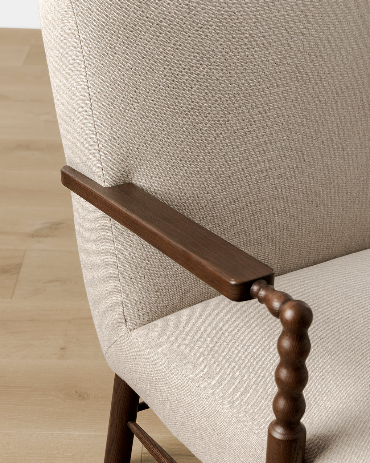 A close-up of the Earnshaw Lounge Chair shows its neutral upholstery and dark English oak armrest with spindle-style support, set on a light wood floor, by McGee & Co.