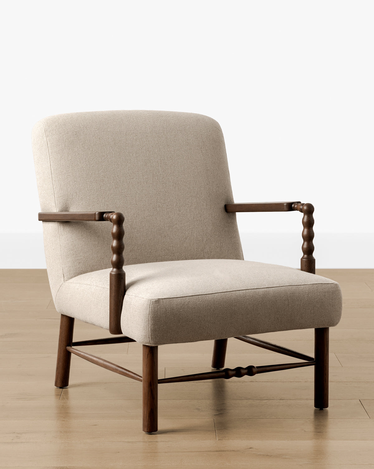 The Earnshaw Lounge Chair has neutral upholstery, wooden legs and armrests in an English oak finish, set on a light wood floor against a plain white wall, by McGee & Co.