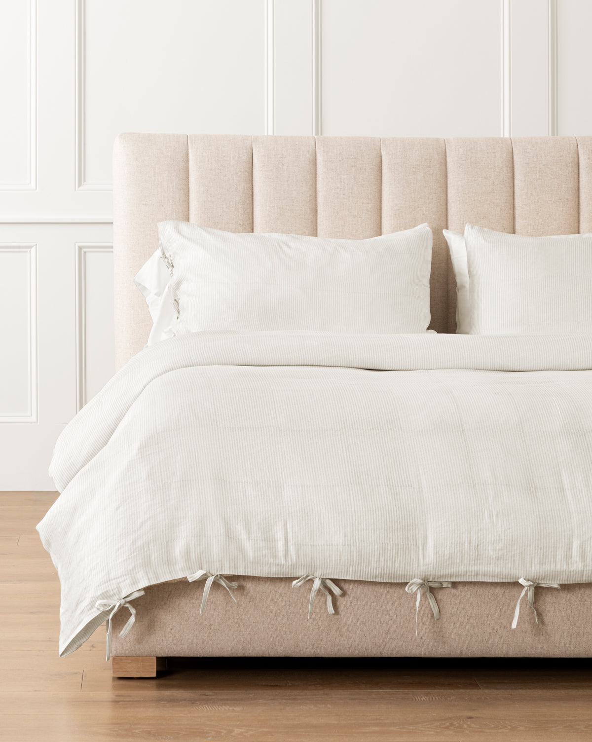 A beige upholstered bed with a tufted headboard is dressed in cream and oatmeal bedding and white pillows. The Easton Striped Duvet Cover, detailed with small ties at the bottom, complements the light wood floor and white-paneled wall.