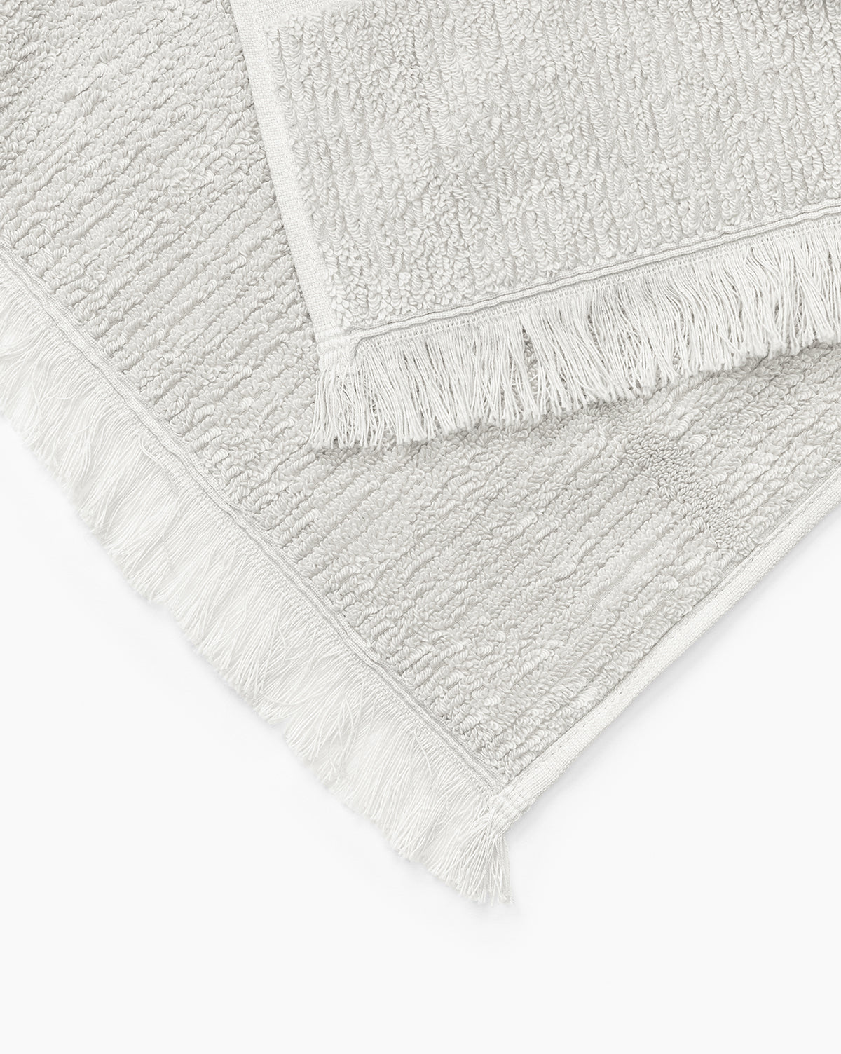 Two light gray textured bath mats from the Edessa Bath Collection, each with fringed edges, are layered together on a white background.