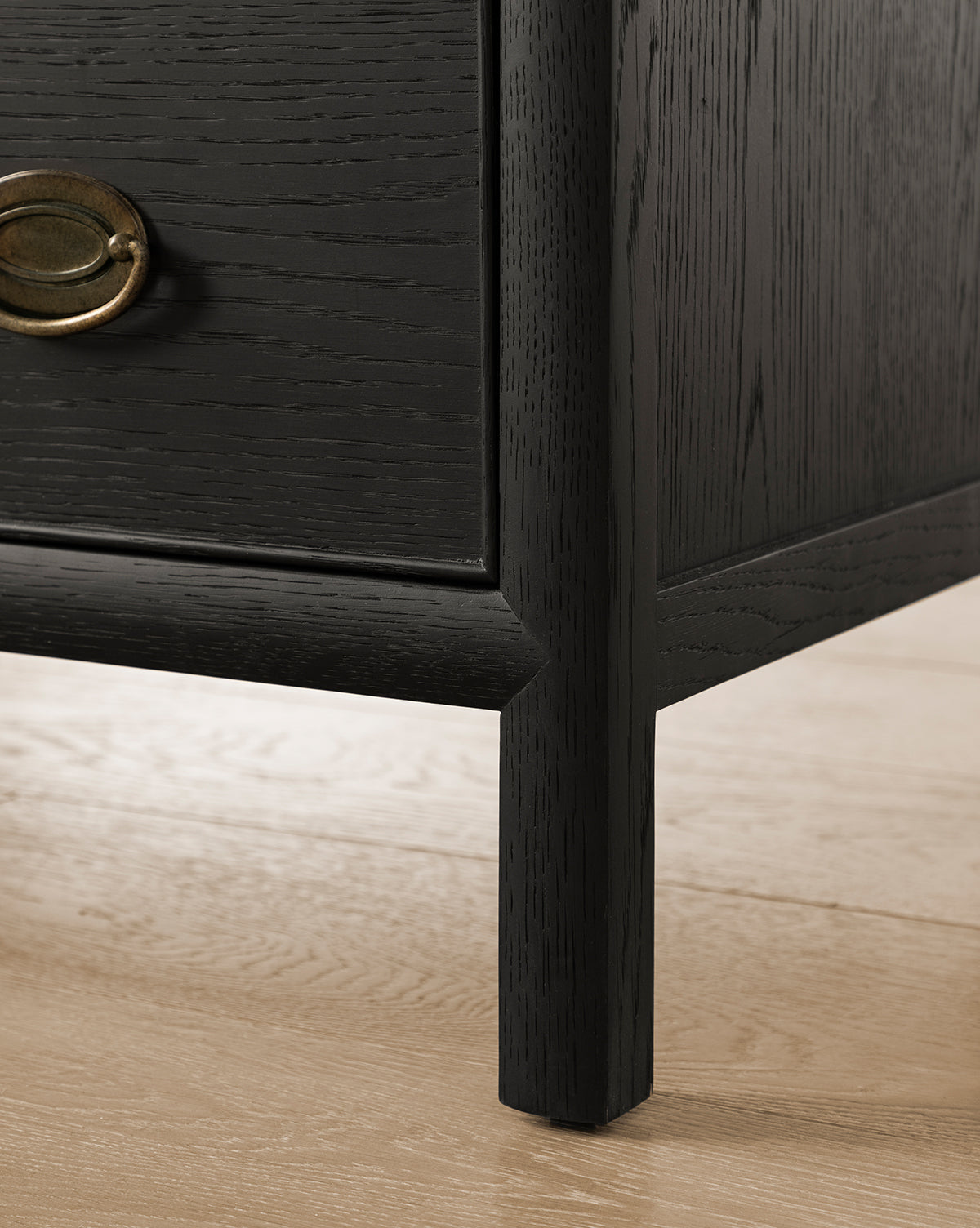 Close-up of the Edward Dresser’s lower corner, showcasing its dark Black Ash finish and brass handle on a light wood floor. The image highlights the textured grain—an elegant bedroom storage solution, by McGee & Co.