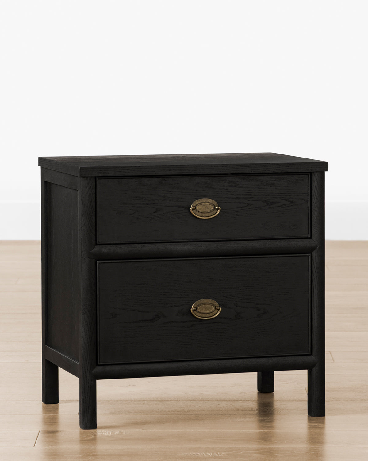 The Edward Nightstand is a black ash bedside table with two drawers featuring brass oval handles, standing on four legs, by McGee & Co.