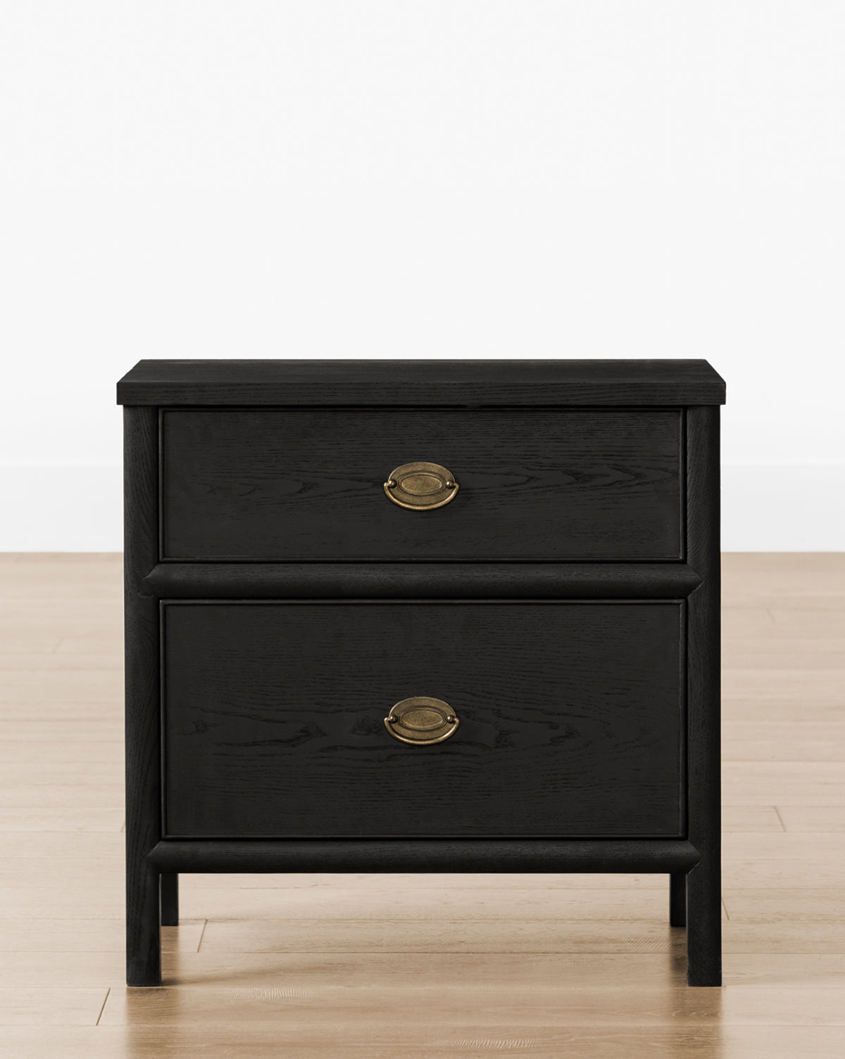 The Edward Nightstand is a black wooden bedside table with two drawers and brass oval handles, shown on a light wood floor against a plain white wall by McGee & Co.