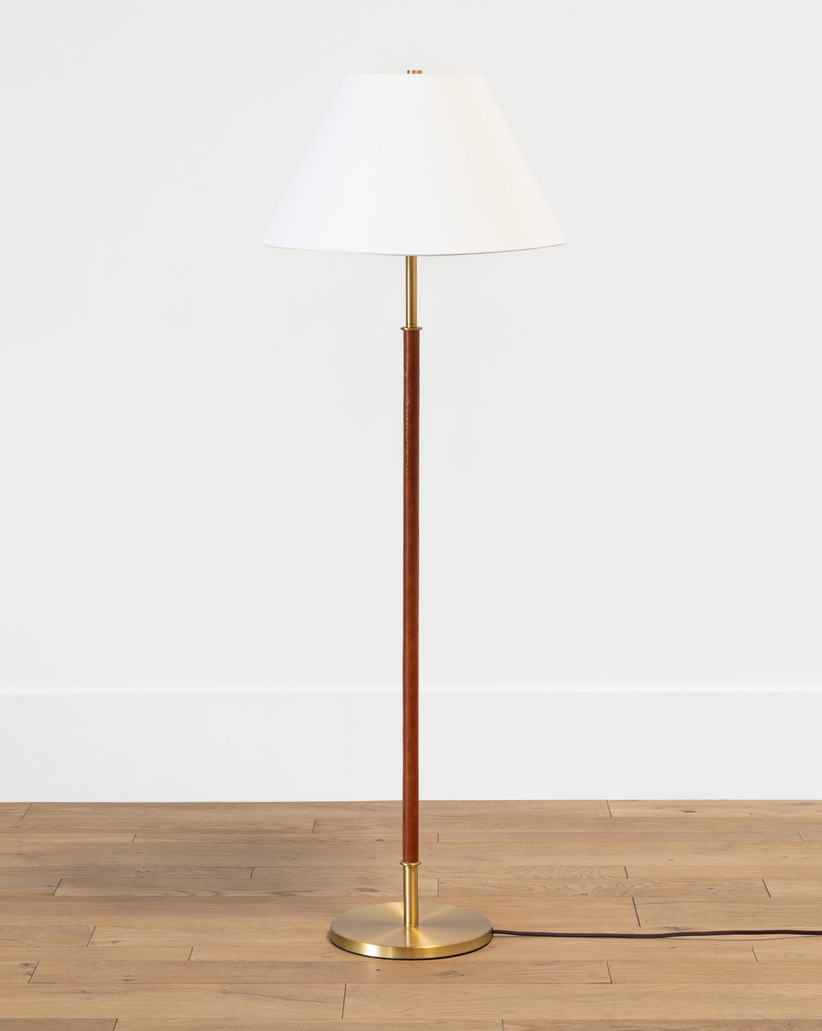 The Elijah Floor Lamp has a white conical shade, slim brass pole, and gold base, shown on a light wood floor with a plain white wall background from McGee & Co.
