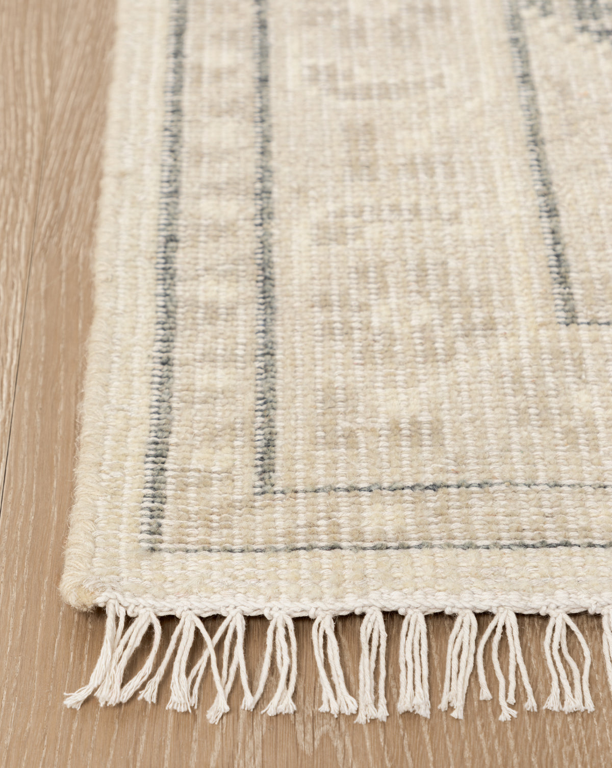 A close-up of the Elin Hand-Knotted Wool Rug, showing its dark border lines and white fringes with artisan details, placed on a light wood floor, by McGee & Co.