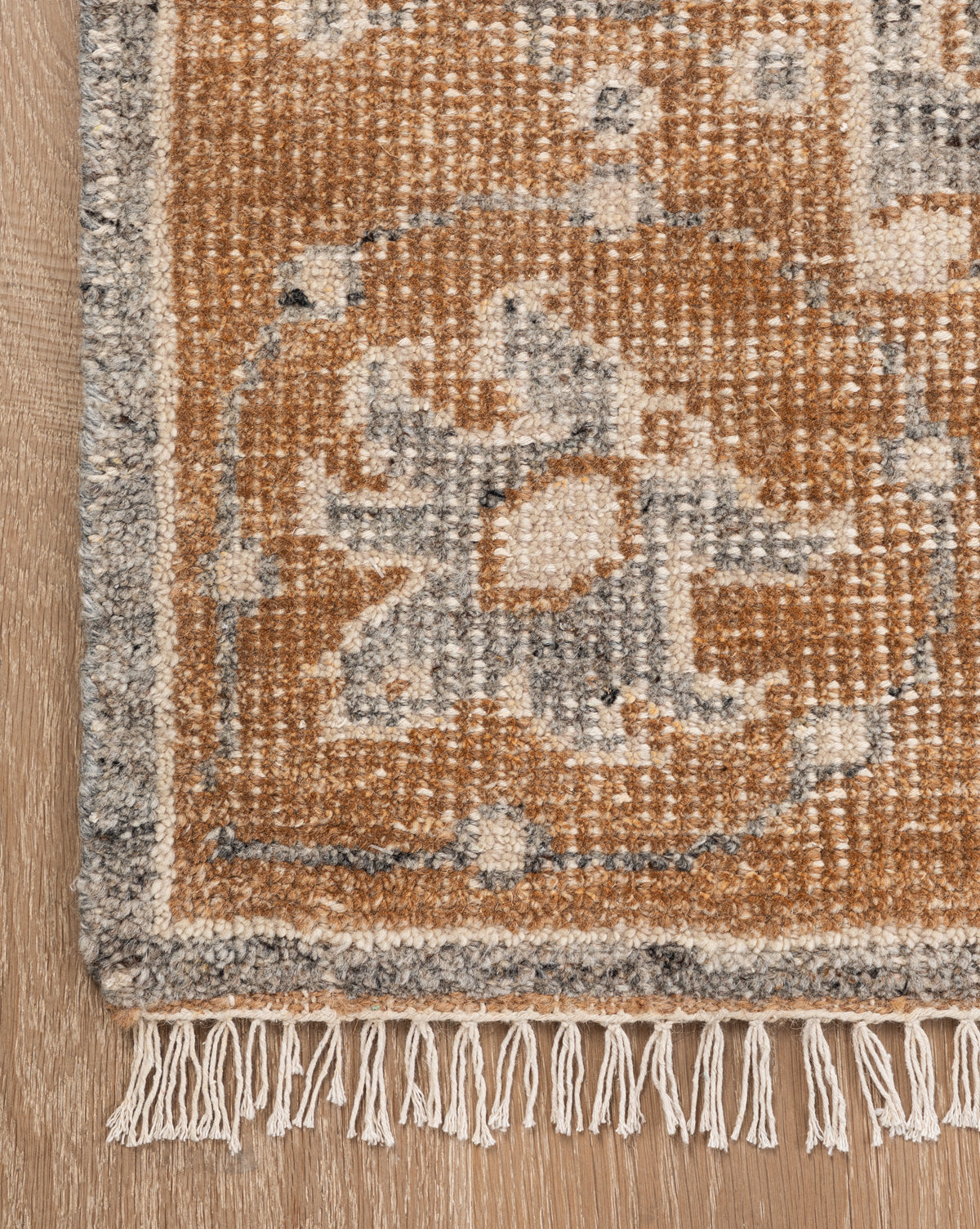 A close-up of the Elison Hand-Knotted Wool Rug corner displays geometric and floral patterns in brown, gray, and cream with short fringe on wood flooring, emphasizing its artisan craftsmanship, by McGee & Co.