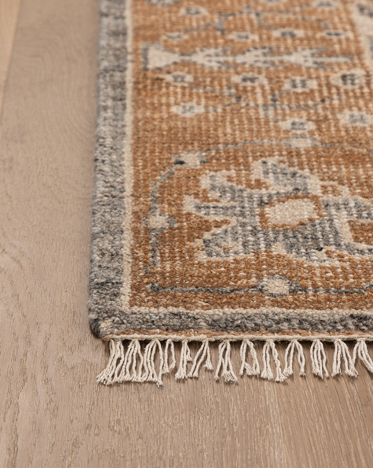 Close-up of the Elison Hand-Knotted Wool Rug’s corner with fringe on light wood flooring. The rug displays earthy brown and gray tones, a geometric pattern, and a decorative gray border, by McGee & Co.