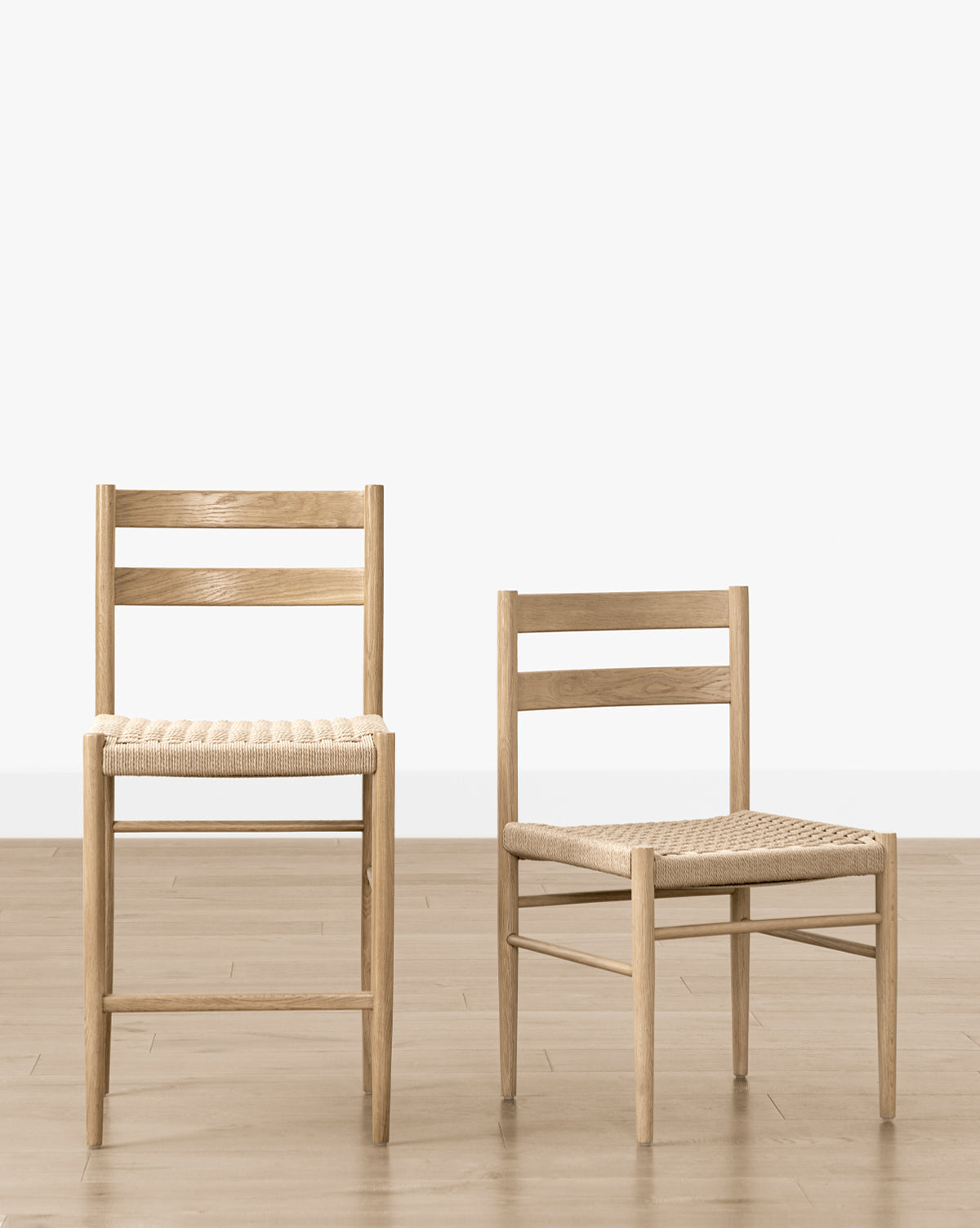 A standard and a shorter wooden chair with woven seats sit side by side on a light wood floor, highlighting minimalist Danish design that perfectly complements the Eloise Woven Counter Stool, by McGee & Co.
