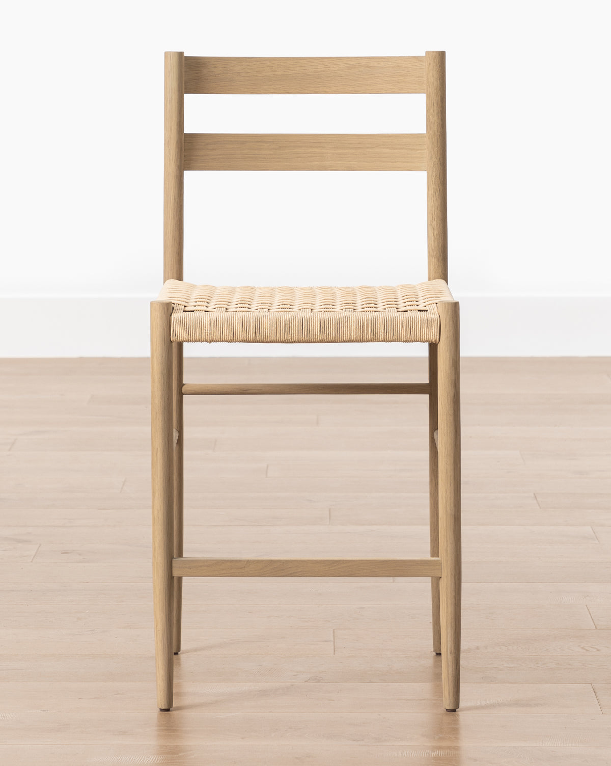 The Eloise Woven Counter Stool, featuring a wooden frame and woven seat with simple straight lines, sits on a light wood floor against a white wall, showcasing classic Danish design, by McGee & Co.