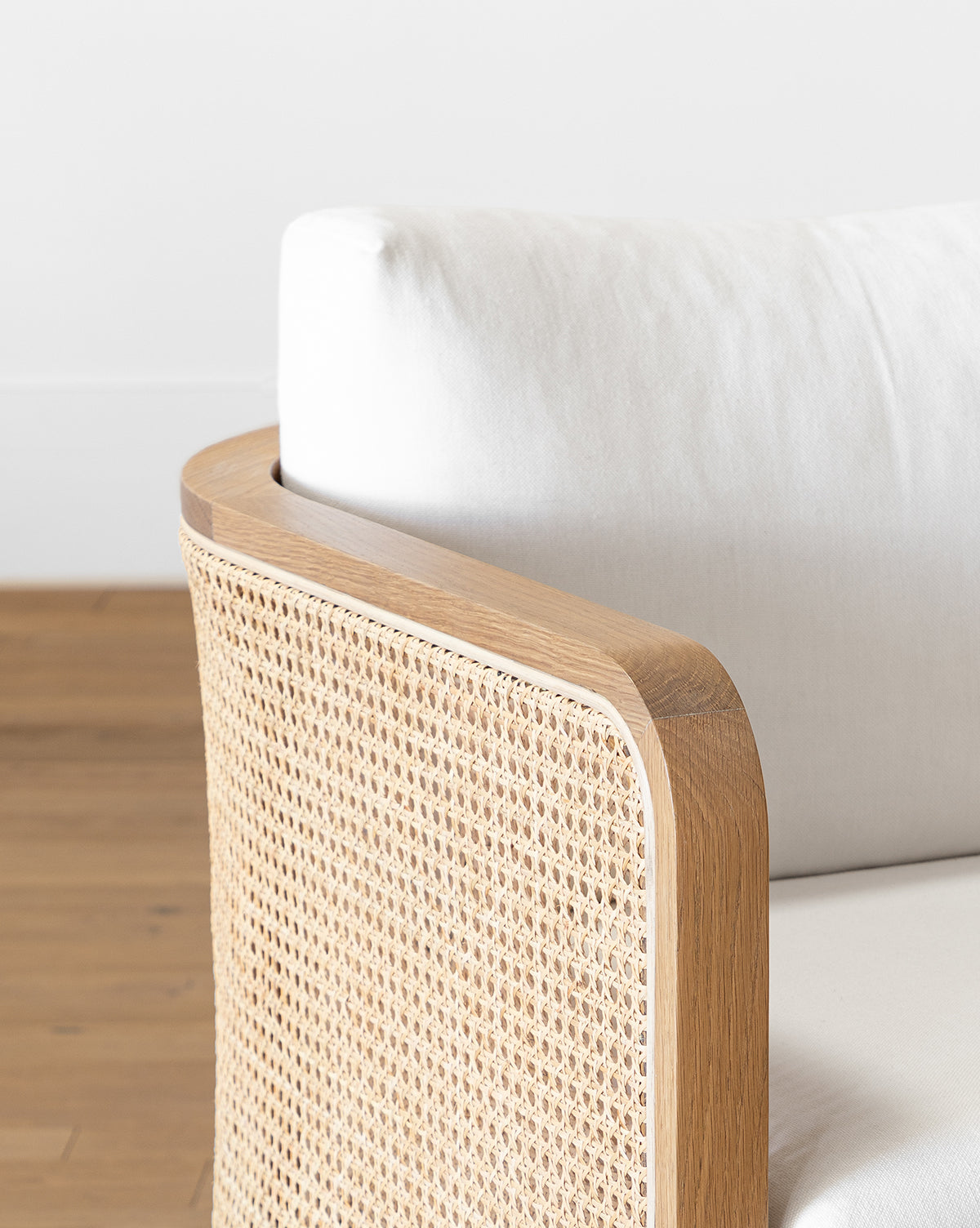 A close-up of the Elowyn Lounge Chair shows its light solid oak frame, natural caning backrest, and white upholstered cushion, placed on a wooden floor with a plain white wall in the background, by McGee & Co.