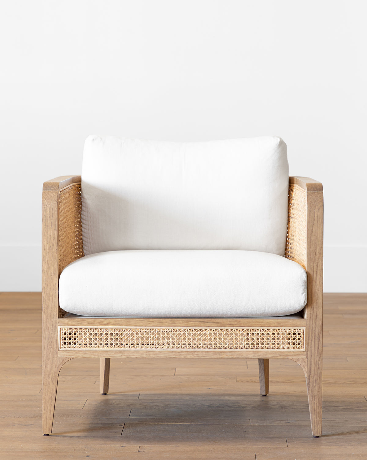 A modern Elowyn Lounge Chair with a solid oak frame, natural caning on the sides and base, and white cushioned seat and backrest sits on a wooden floor against a plain white wall by McGee & Co.