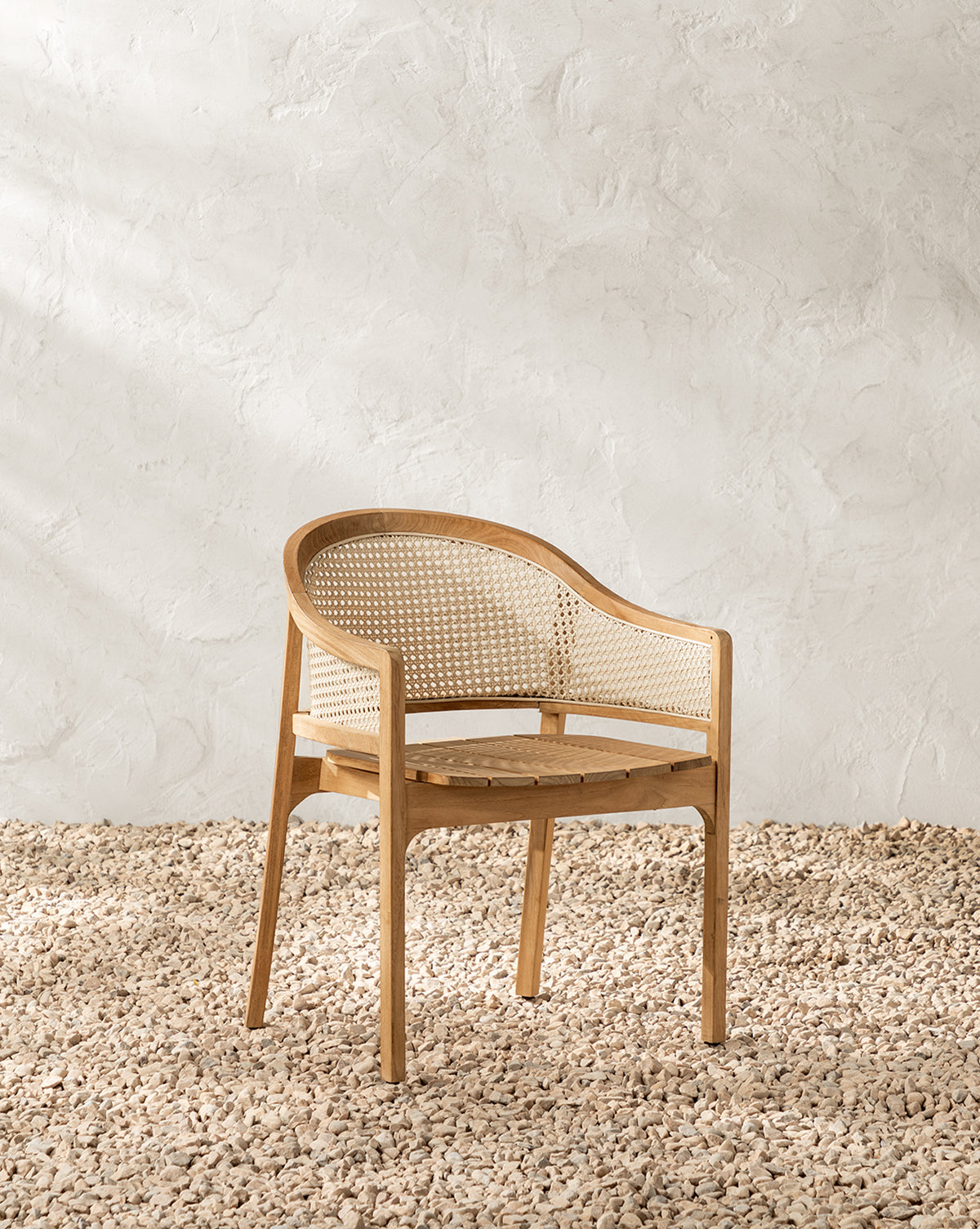 The Elowyn Outdoor Dining Chair, featuring a curved backrest and cane woven seat, sits on light-colored pebbles with a textured white wall as the backdrop, by McGee & Co.