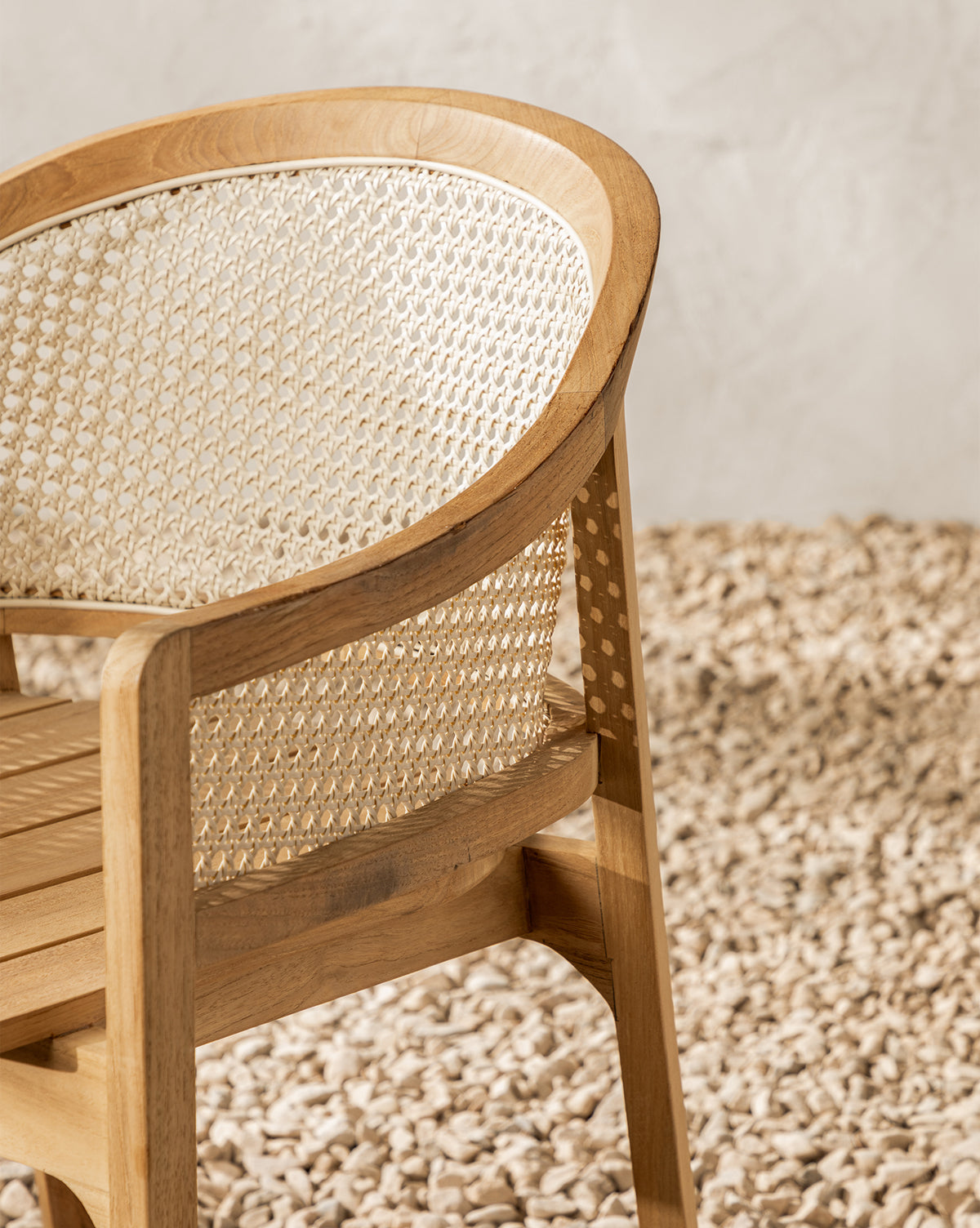 A close-up of the Elowyn Outdoor Dining Chair shows its curved cane backrest and teak finish, set on small, light-colored stones with a subtly patterned light wall in the background, by McGee & Co.