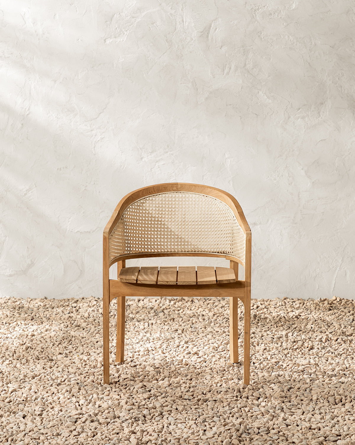 The Elowyn Outdoor Dining Chair, featuring a teak frame and woven cane backrest, sits on a textured pebble floor against a white textured wall in a minimalistic setting by McGee & Co.