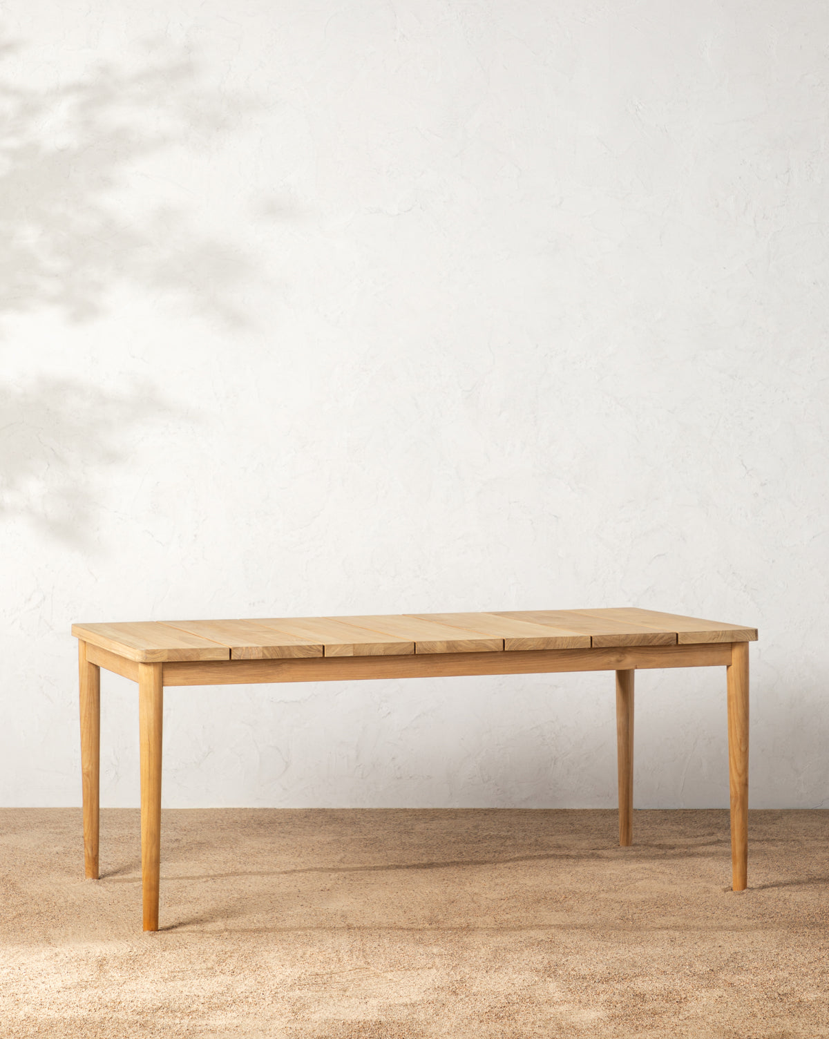The Elowyn Outdoor Dining Table, a simple rectangular teak piece with four legs, stands on a light brown carpet by a textured white wall, with soft foliage shadows cast on the left side of the wall, by McGee & Co.