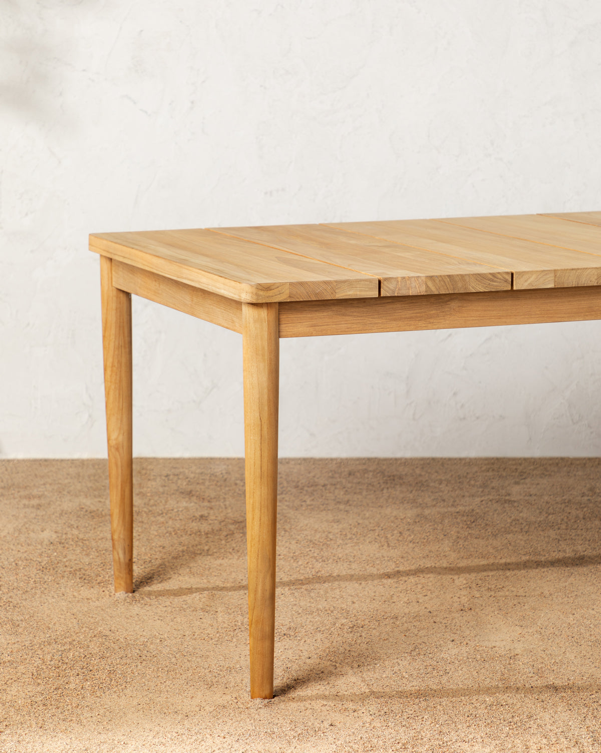 The Elowyn Outdoor Dining Table, crafted from light teak wood with tapered legs, stands on a textured beige floor against an off-white wall—ideal for all-weather outdoor dining, by McGee & Co.