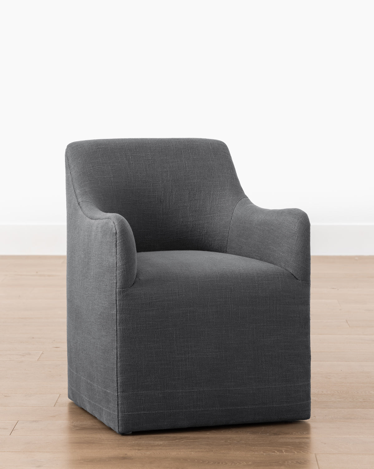 The Elton Dining Chair, featuring a modern charcoal gray linen-blend upholstery with curved back and arms, sits on a light wood floor against a plain white wall—ideal for any dining space, by McGee & Co.