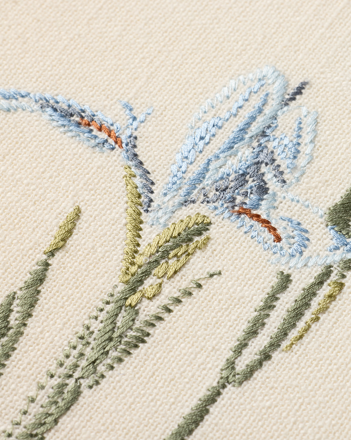 Close-up of the Embroidered Blue Iris features delicate cotton embroidery on beige fabric, with blue iris flowers, green stems and leaves, and subtle orange accents. The textured threads enhance this botanical wall art, by McGee & Co.