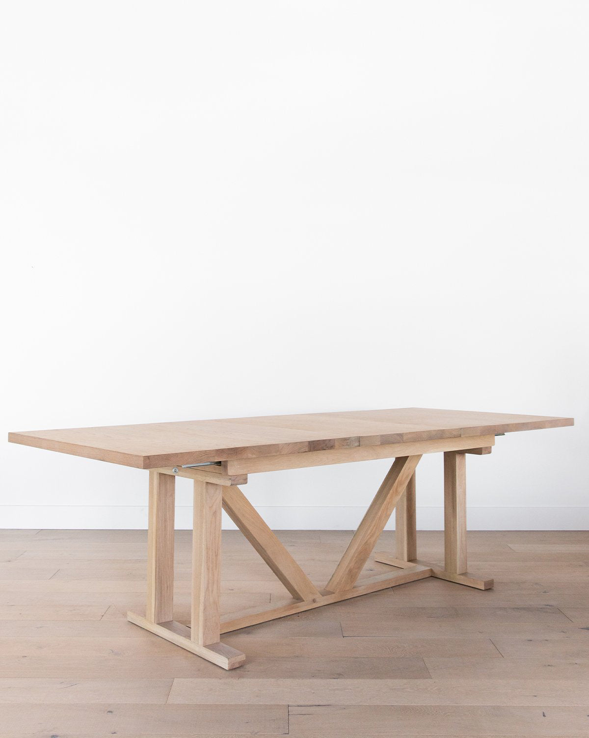 The Emory Extension Dining Table, featuring a thick solid oak top and sturdy trestle-style base, sits on a light wood floor against a plain white wall, by McGee & Co.