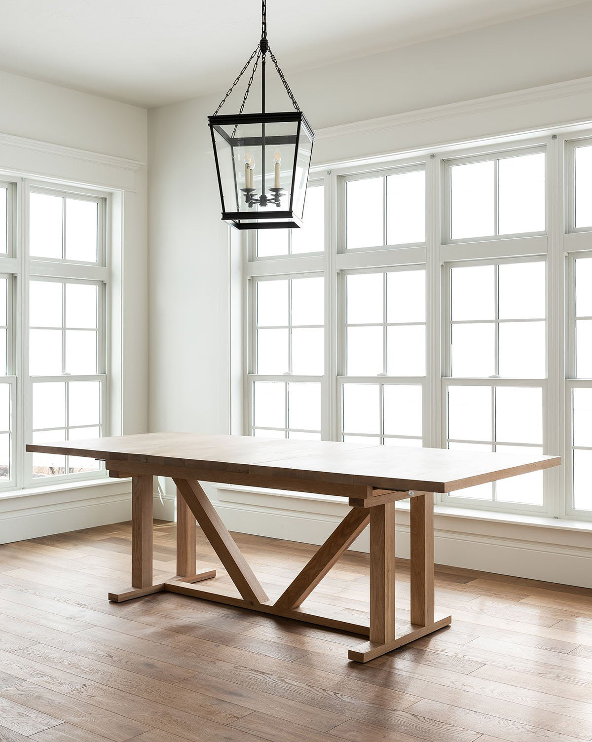 A minimalist room with large, white-framed windows features the Emory Extension Dining Table with a trestle base, paired with a black metal lantern pendant light hanging above on a wooden floor, by McGee & Co.